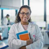 Medical Science Laboratory: Beautiful Smart Young Black Scientist Wearing White Coat and Glasses, Holds Test Books, Smiles Looking at Camera. Diverse Team of Specialists. Medium Portrait Shot