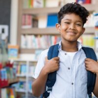 Portrait of smiling hispanic boy looking at camera. Young elementary schoolboy carrying backpack and standing in library at school. Cheerful middle eastern child standing with library background.