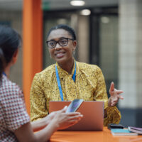 Two female teachers sitting at a table, having a discussion in the lobby area in the school they work at in Gateshead, North East England. They are talking about school issues together while using a laptop.